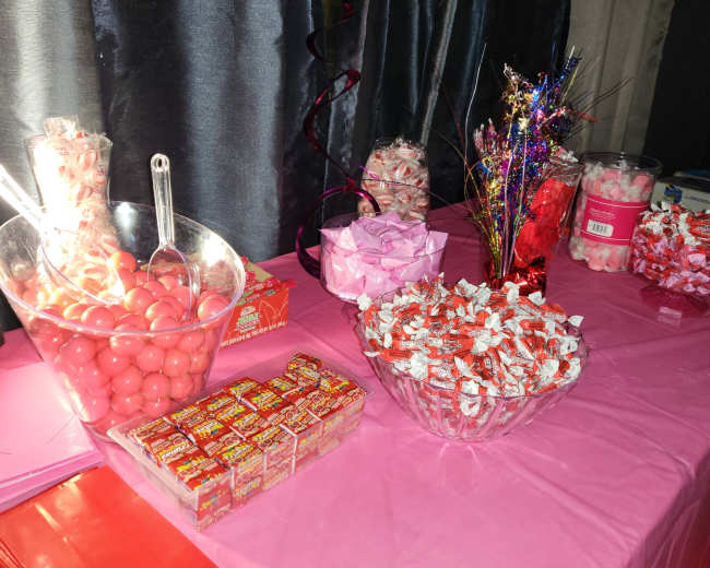 A table covered in pink cloth displays various containers filled with colorful candies, including pink gumballs, festive decorations, and assorted wrapped treats.