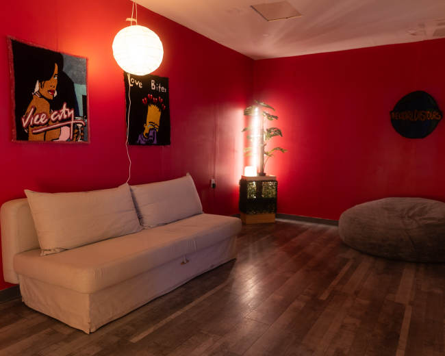 The image shows a room with bright red walls, featuring a white sofa, a beanbag chair, and artwork on the walls, illuminated by soft lighting.