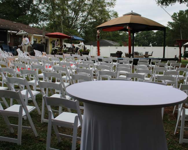 The image shows a neatly arranged outdoor event space with white chairs set up in rows, a central round table, and a large gazebo in the background.