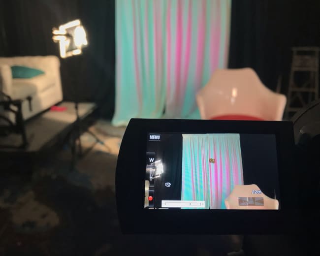 A camera viewing a softly lit studio backdrop with curtains and seating arrangements set up for a video shoot.