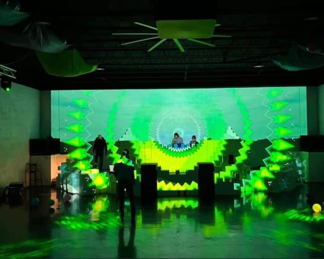 A performance area features a vibrant green digital visual display with two DJs in the background.