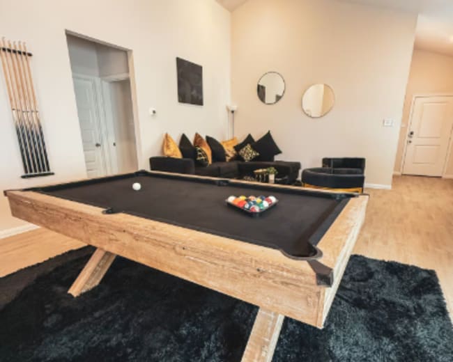 A wooden pool table with a black felt surface is positioned on a black rug in a brightly lit room featuring a couch and decorative mirrors.