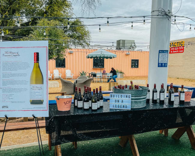 A display table featuring multiple bottles of wine, with a sign and a container of ice, is set up in a outdoor venue with string lights overhead.