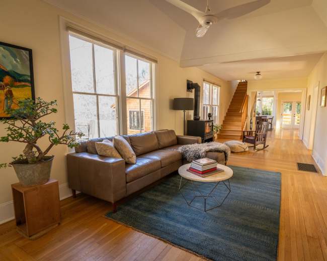 The image shows a bright, open living room with a brown leather sofa, a round table with books, and wooden flooring leading to a staircase.