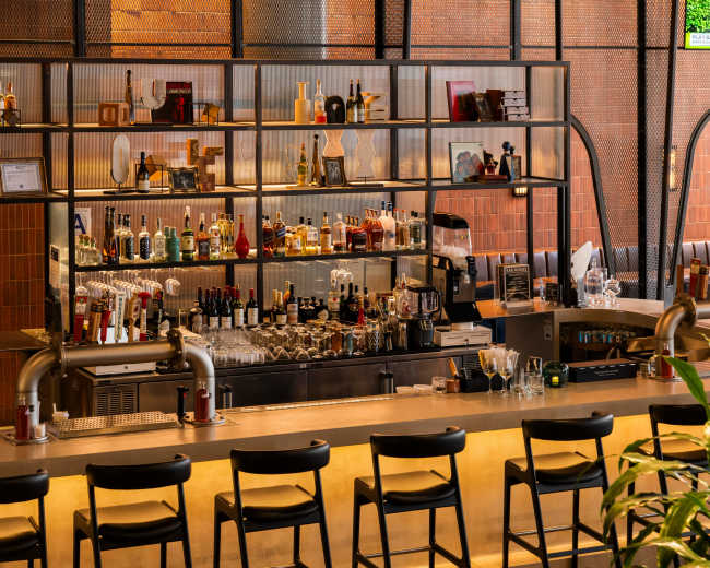 The image shows a modern bar with a well-stocked shelf of various liquor bottles and a long countertop lined with black chairs.