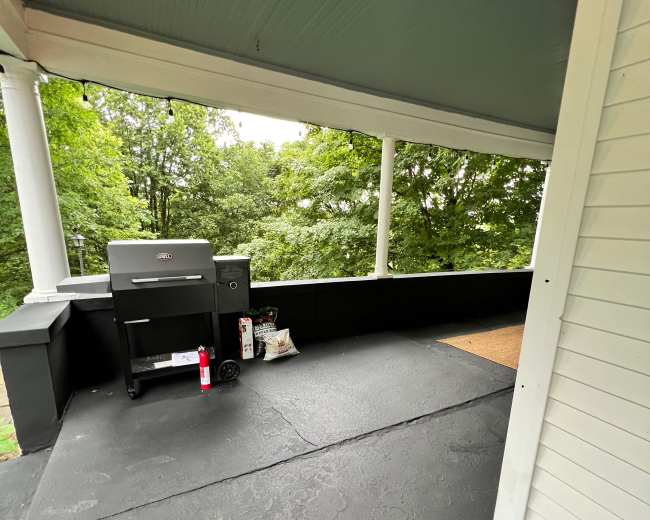 A covered porch with a grill and various items positioned near a railing overlooking a green landscape.