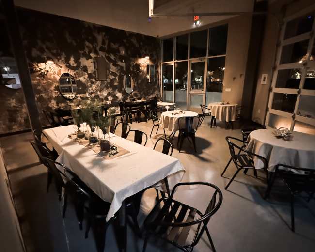 The image shows a dimly lit restaurant interior with several tables set for dining, featuring simple tablecloths and black chairs.