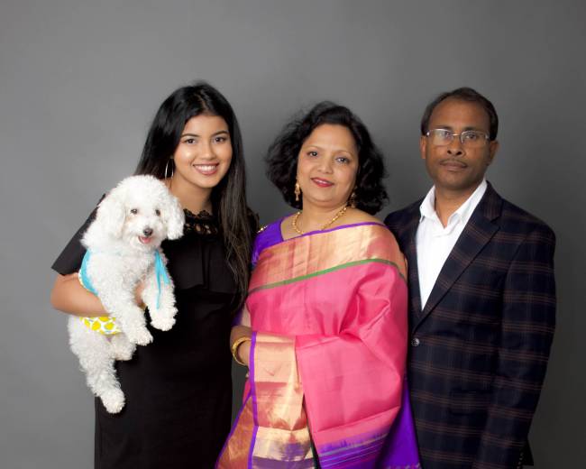 A woman in a black dress holds a small dog while standing next to a woman in a colorful sari and a man in a checkered suit.