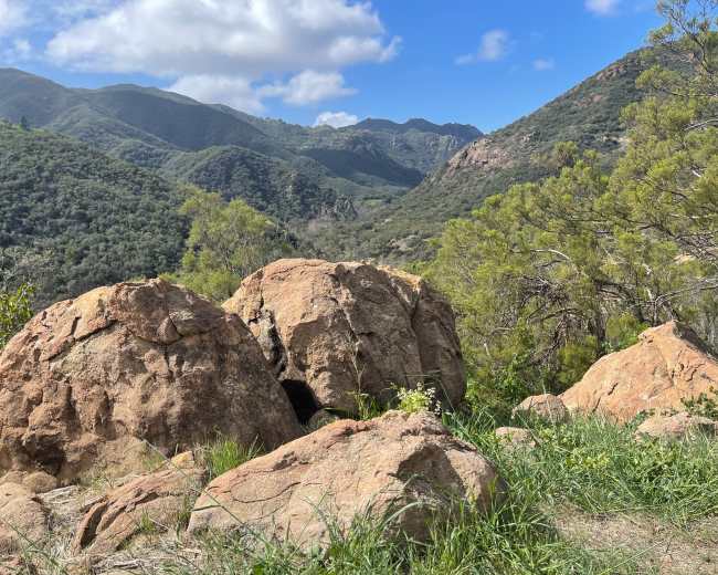 A cluster of large boulders sits at the forefront with rolling green hills and mountains in the background under a partly cloudy sky.