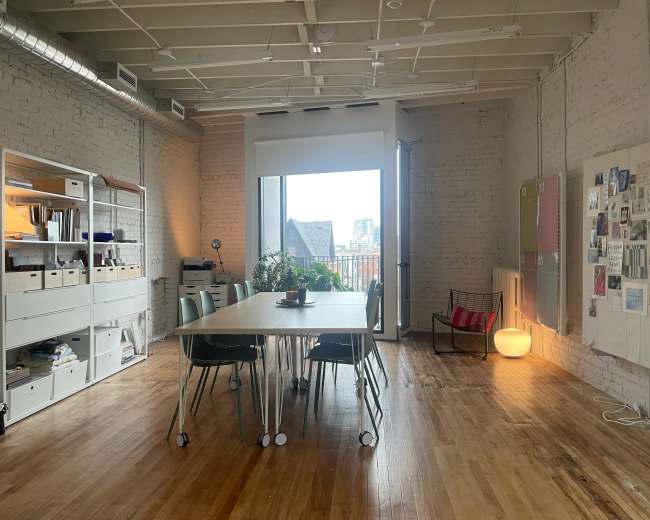 A large table is surrounded by chairs in a sunny, minimalist room featuring exposed brick walls and a window showing a city skyline.