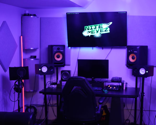 The image shows a home recording studio with a computer desk, speakers, and soundproofing materials, illuminated by purple lighting.