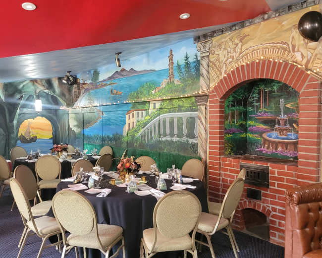 The image shows a dining area with round tables set for a meal, surrounded by a mural depicting a scenic landscape and a decorative brick archway.