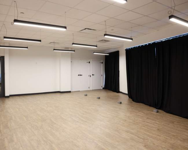 The image shows a spacious room with a wooden floor, overhead lighting, and black curtains on one side.