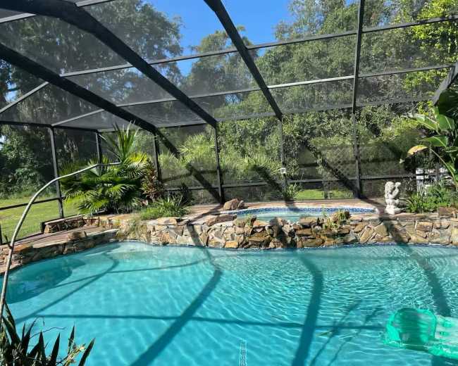 The image shows a swimming pool enclosed by a mesh screen, surrounded by greenery and a stone wall.