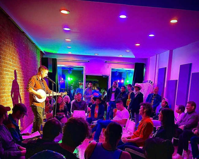A musician performs with a banjo in front of an audience seated in a brightly lit room with colorful stage lighting.