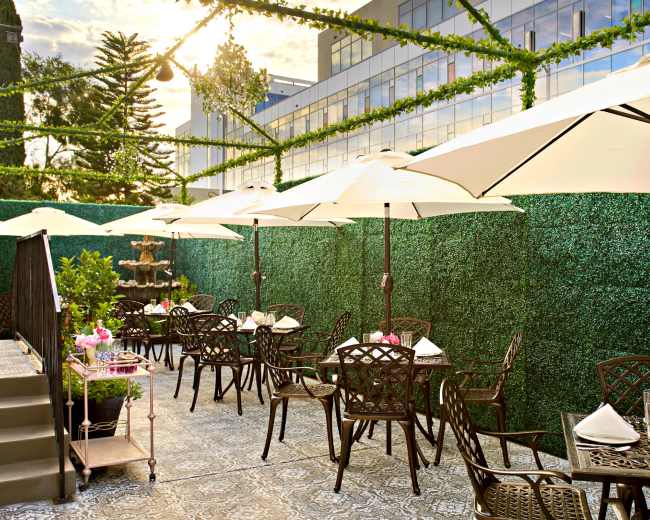 An outdoor dining area with tables set under large umbrellas, surrounded by lush green walls and a fountain in the background.