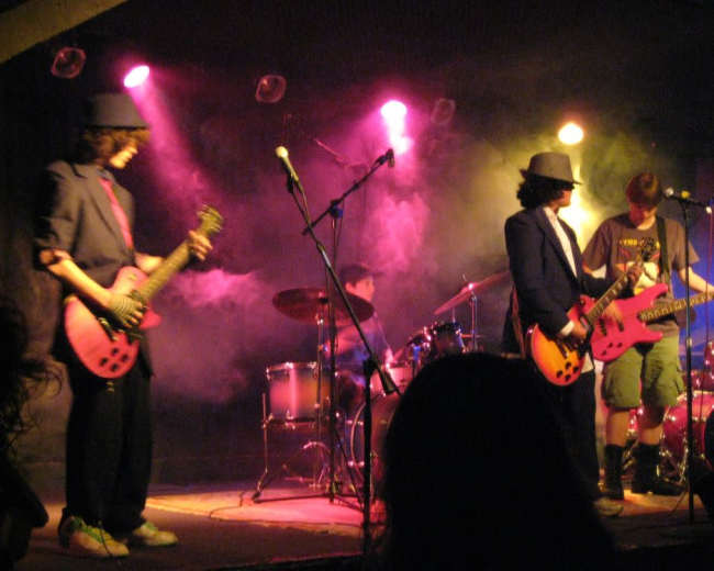 A band performs on stage with electric guitars and a drum set, surrounded by colorful stage lights and fog.