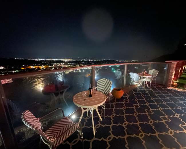 A dimly lit terrace features two chairs and a small table overlooking a city skyline at night.