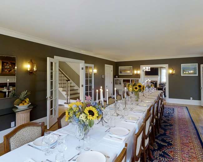 A long dining table is set with glasses and floral arrangements in a room featuring a fireplace and large windows.