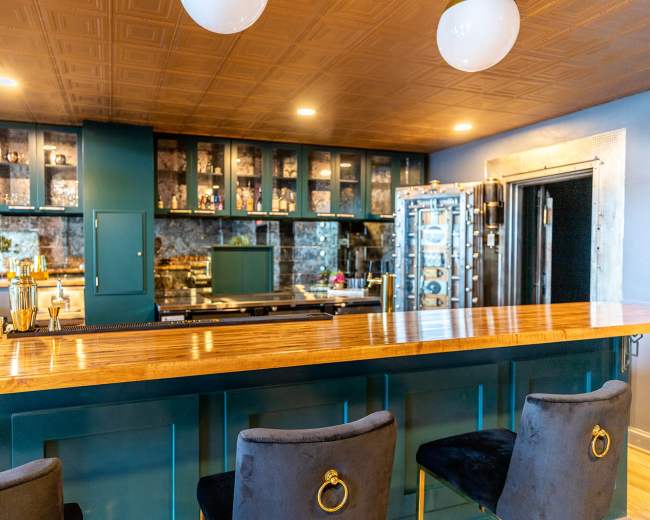 The image shows a modern bar area with a wooden countertop, black upholstered stools, and a wall of glass-front cabinets displaying various items.
