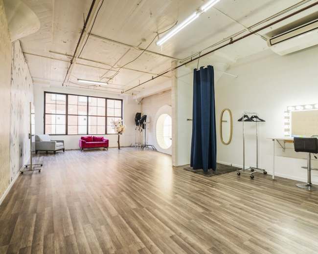 An open studio space with wooden flooring, a decorative wall, a large window, a seating area, and a makeup station with a lit mirror.