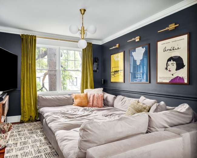 The room features a cozy sectional sofa, decorative posters on the walls, large windows with yellow curtains, and a mounted television.