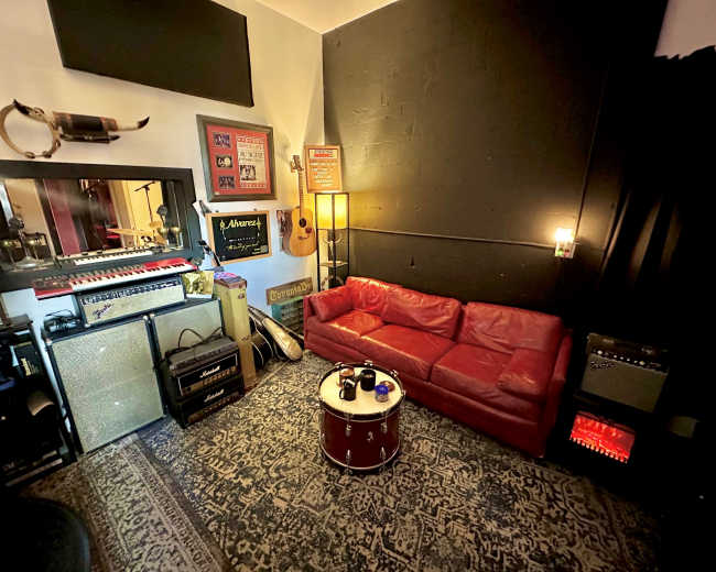 The image shows a cozy music studio featuring a red leather couch, musical instruments, amplifiers, and sound equipment arranged against a black wall.
