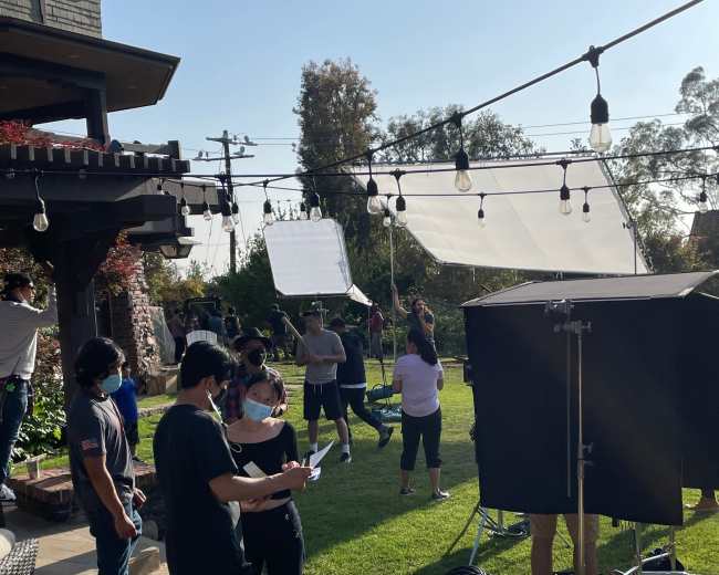 A group of people is working on a film set in a backyard, setting up lights and equipment under a clear sky.