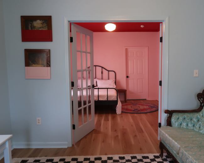 A light blue room featuring a green upholstered sofa, framed artwork on the wall, and an open doorway leading to a bedroom with a bed and a closed door.