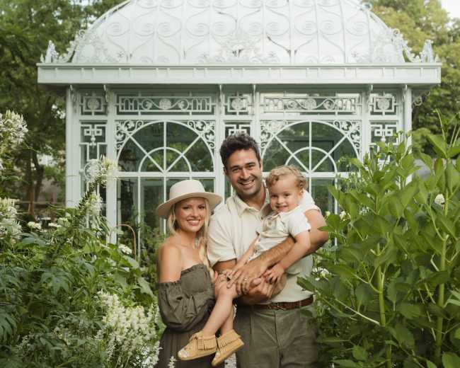 A family of three stands in front of a glass greenhouse surrounded by lush greenery and flowering plants.
