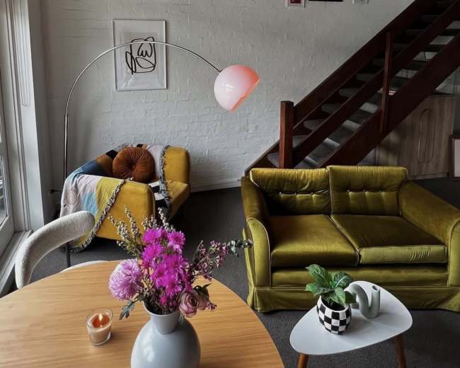 A cozy living room features a yellow couch, a patterned armchair, a round dining table with a vase of flowers, and artwork displayed on the walls.