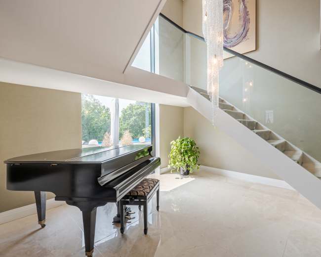 A black grand piano is positioned near a glass staircase in a well-lit foyer with a large window overlooking greenery.