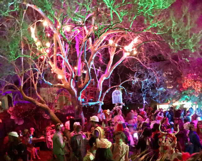 A lively gathering takes place at night under a colorful illuminated tree, with guests wearing various costumes mingling in a festive atmosphere.