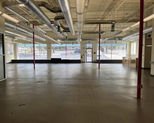 An empty commercial space with large windows, exposed ductwork, and a concrete floor.
