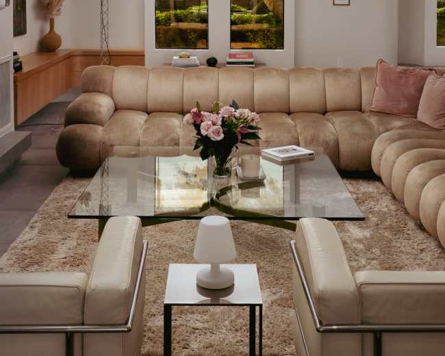 A modern living room features a large, curved beige sofa, a glass coffee table on a plush rug, and potted plants visible through the windows.
