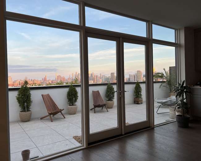 The image shows a modern indoor space with large glass doors leading to a terrace that overlooks a city skyline.