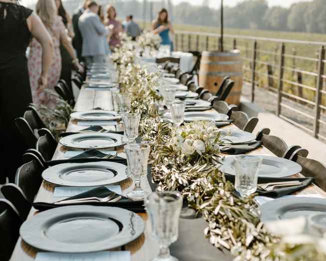 A long outdoor table is set for a gathering, adorned with black plates, glassware, and a green garland centerpiece.