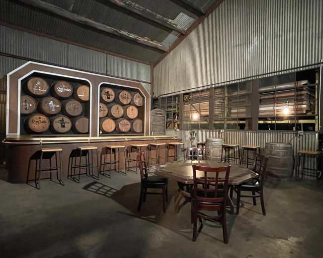 A spacious, dimly lit warehouse featuring a bar area with wooden stools and a round table surrounded by barrels and a large wall of decorative barrels.