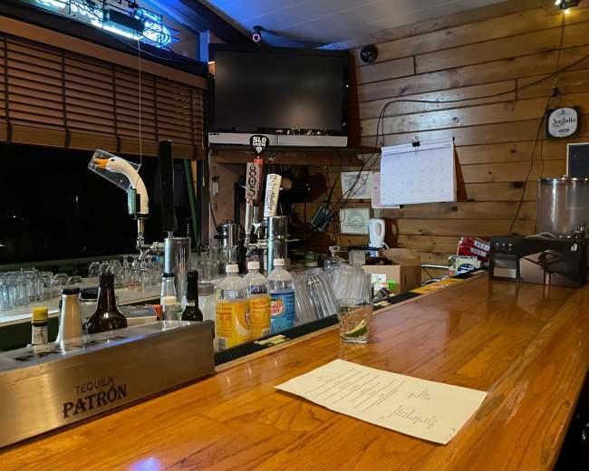 A bar counter with a variety of liquor bottles, glasses, and a menu rests under wooden panels illuminated by a neon sign.
