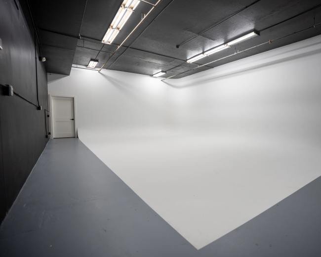 A spacious, empty studio with white walls and a gray floor, featuring a single door on one side.