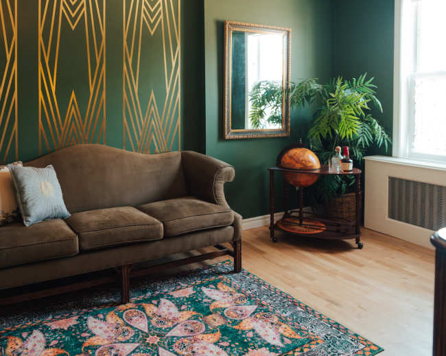 The image shows a living room featuring a brown sofa, a decorative area rug, a potted plant, a globe on a cart, and a mirror on a green accent wall with gold geometric patterns.