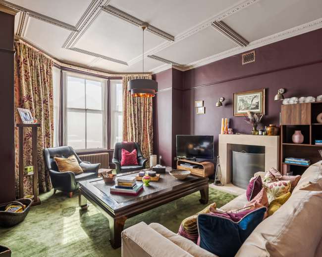 The image shows a well-furnished living room featuring a coffee table, seating area with a sofa and chairs, and a large window with patterned curtains.