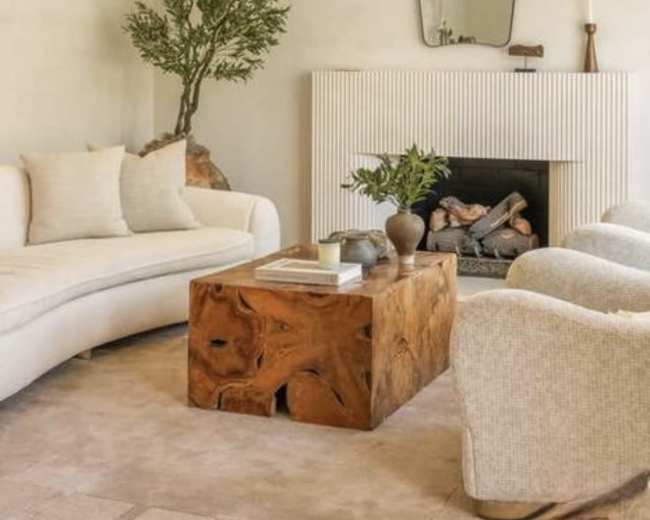 The image depicts a minimalist living room featuring a curved white sofa, a wooden coffee table, and two textured armchairs, with a fireplace and a large mirror on the wall.