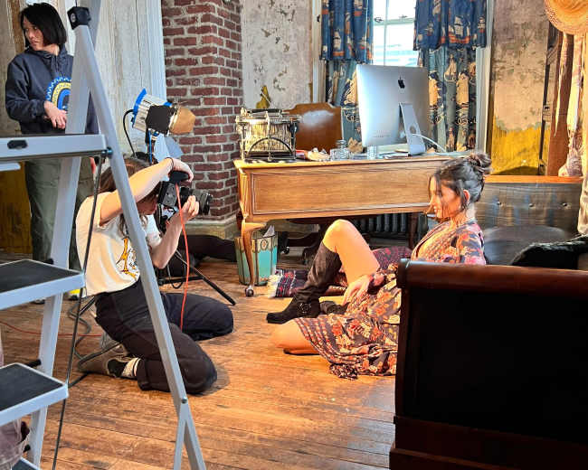 A photographer captures an image of a model sitting on the floor in a vintage-styled room while another person assists in the background.