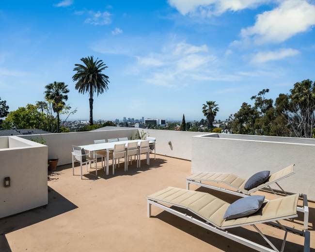 The image shows a rooftop terrace with white patio furniture, including a dining table and lounge chairs, overlooking a cityscape with palm trees in the background.