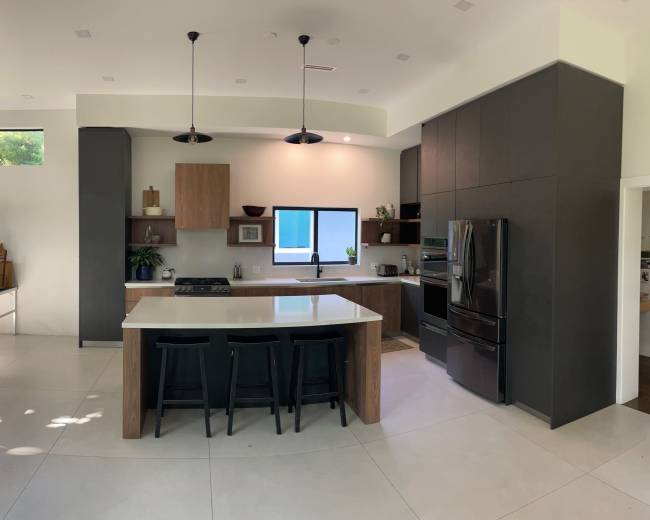 The image shows a modern kitchen featuring a central island with seating, dark cabinetry, stainless steel appliances, and a large window that allows natural light to enter the space.