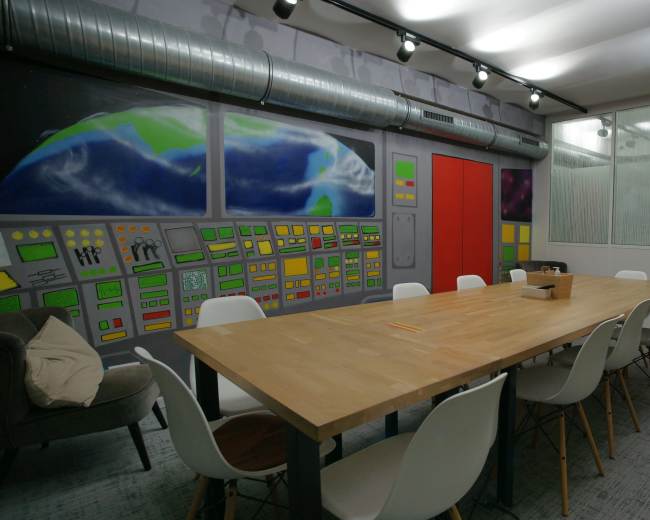 The image shows a modern conference room with a large wooden table surrounded by white chairs, featuring colorful murals on the walls and a glass-paneled area.