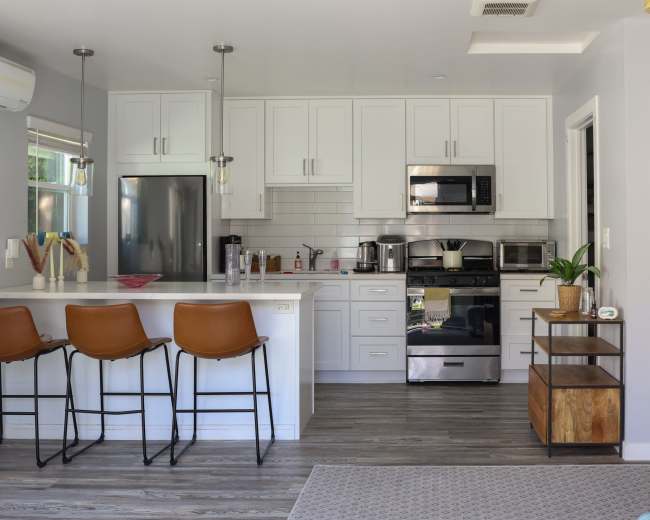The image shows a modern kitchen with white cabinets, stainless steel appliances, and a countertop with three brown bar stools.