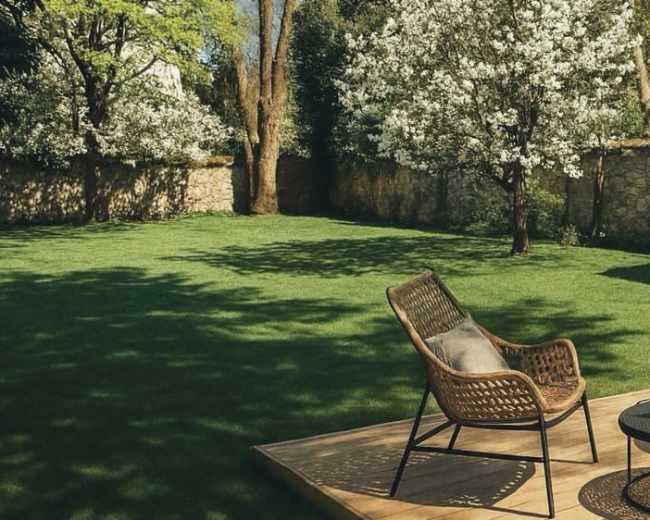 The image shows a backyard with a well-manicured lawn, two flowering trees, a stone wall, and a wooden deck with a chair and small table.