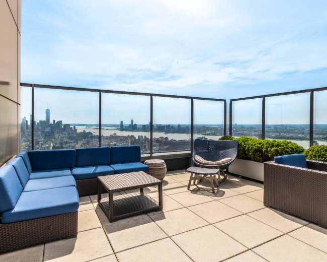 The image shows a rooftop terrace with blue seating, a small table, and a view of a city skyline and river.